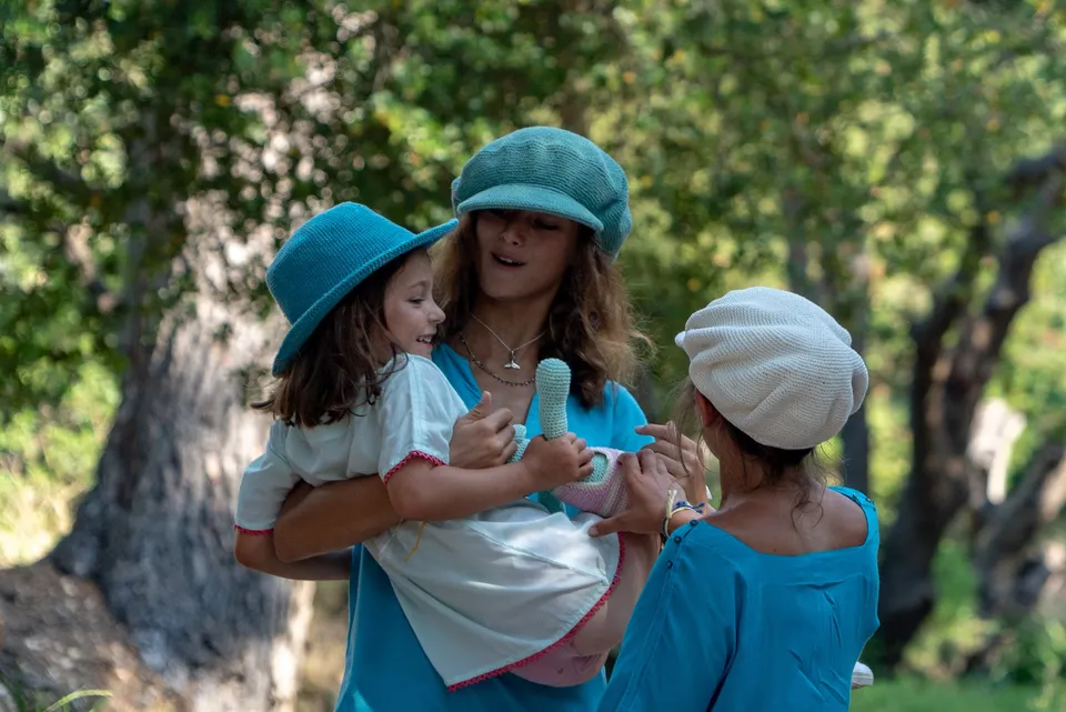 Deux femmes en tuniques turquoise et chapeaux crochet portent une fillette souriante dans le maquis de Porquerolles.