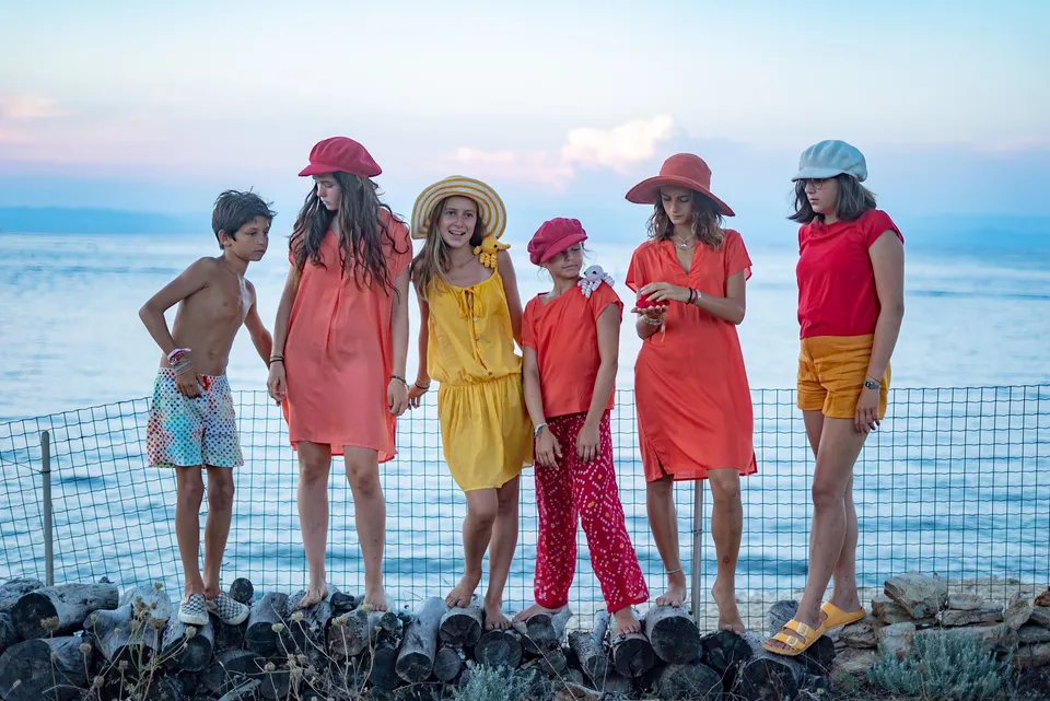 Groupe d'enfants en robes rouge, orange et jaune sur des rondins face à la mer au crépuscule à Porquerolles.