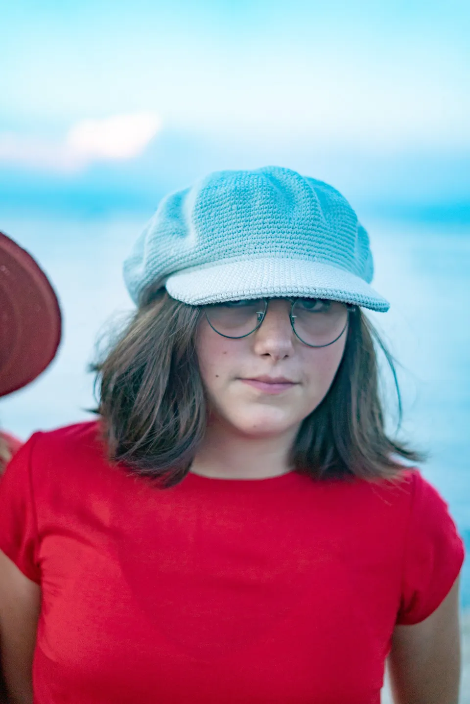 Portrait d'une jeune fille en t-shirt rouge, casquette crochet bleu-gris et lunettes, mer floue à Porquerolles.