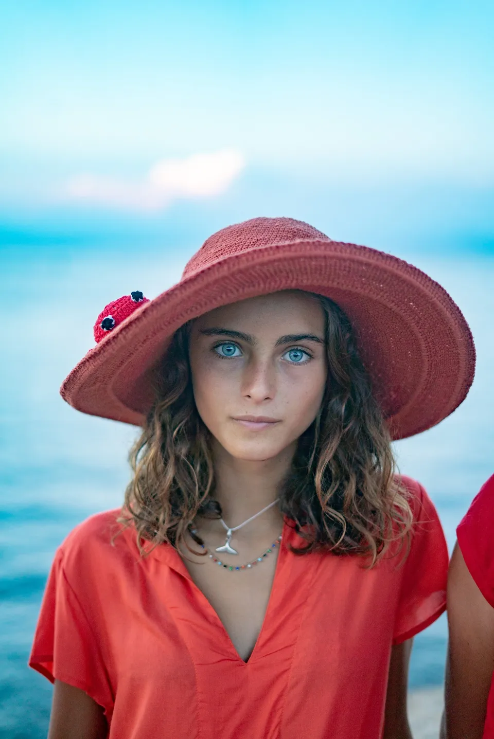 Enfant en blouse rouge-orangé et chapeau rouge à coccinelle crochetée, mer bleue floue derrière à Porquerolles.