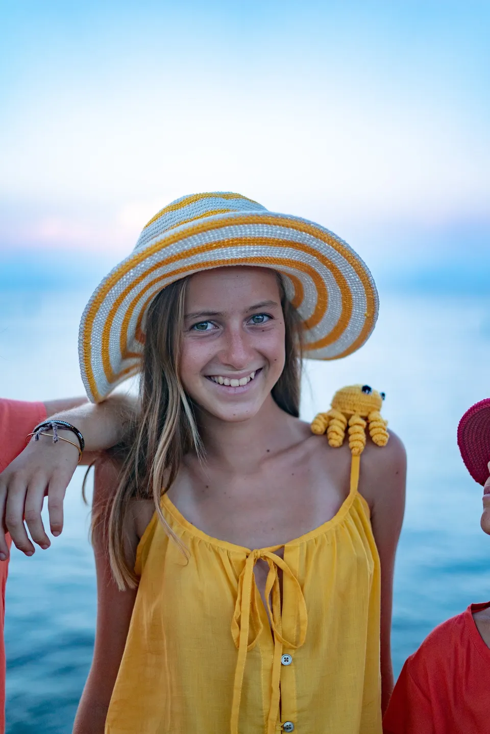Enfant souriante en robe jaune et chapeau rayé, pieuvre de crochet sur l'épaule face à la mer à Porquerolles.