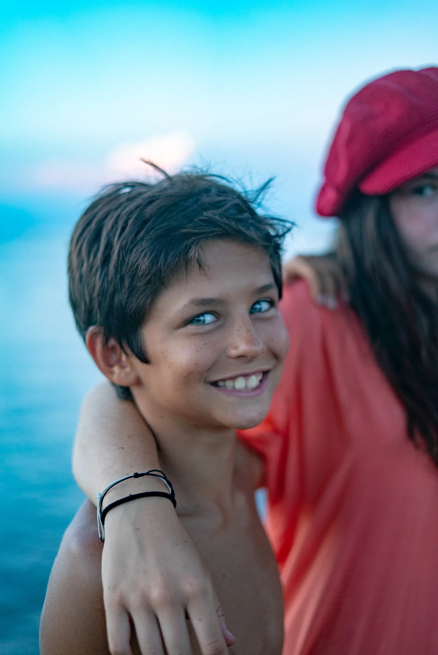 Garçon torse nu souriant serré par une fillette en haut corail et casquette rouge, mer floue derrière à Porquerolles.