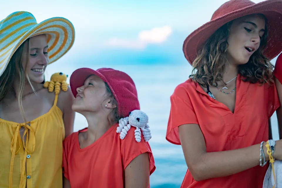 Trois fillettes en jaune et rouge-orange, chapeaux et pieuvres crochetées, face à la mer à Porquerolles.