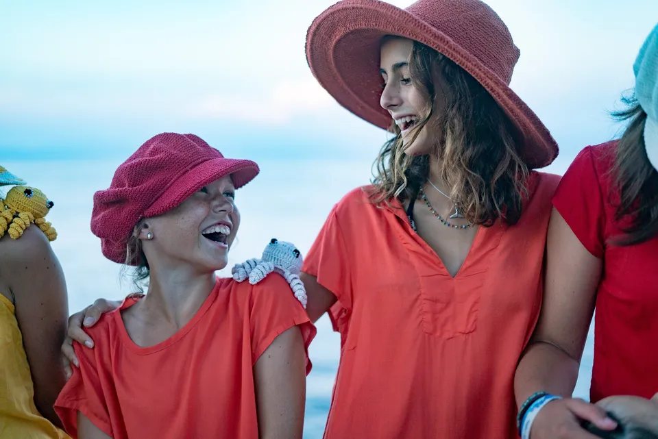 Deux jeunes filles riant en tuniques rouges, pieuvres crochetées sur l'épaule, mer derrière à Porquerolles.