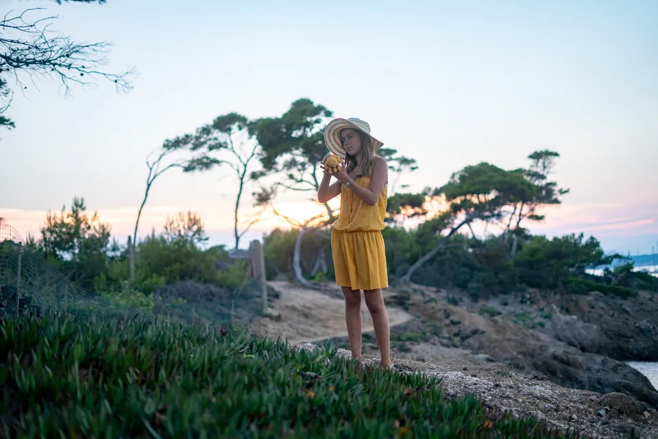 Jeune fille en robe jaune et chapeau rayé sur un sentier côtier, pins et coucher de soleil à Porquerolles.