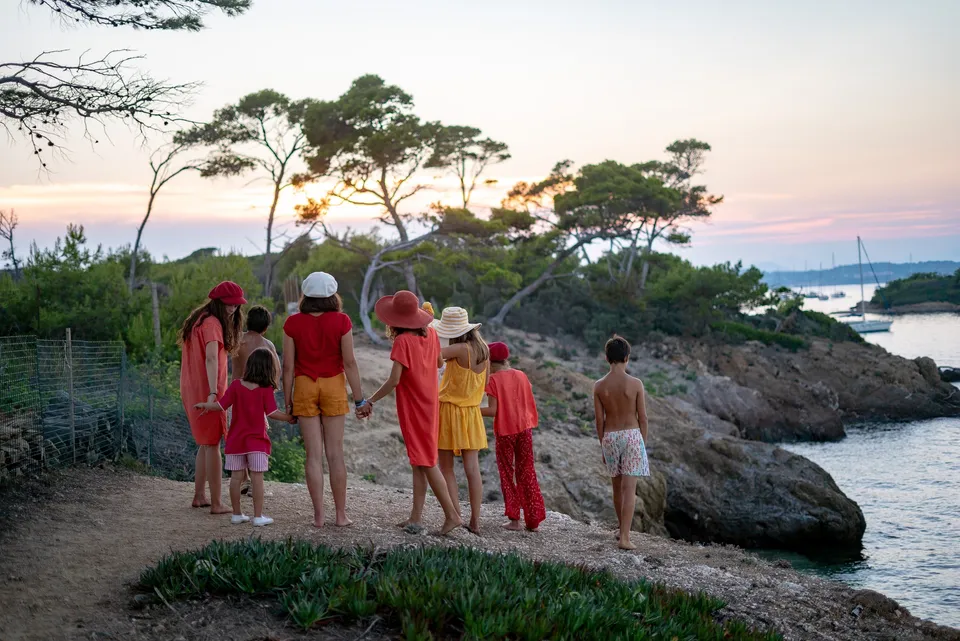Enfants de dos en tenues rouge et orange sur un sentier rocheux face à la baie au couchant à Porquerolles.