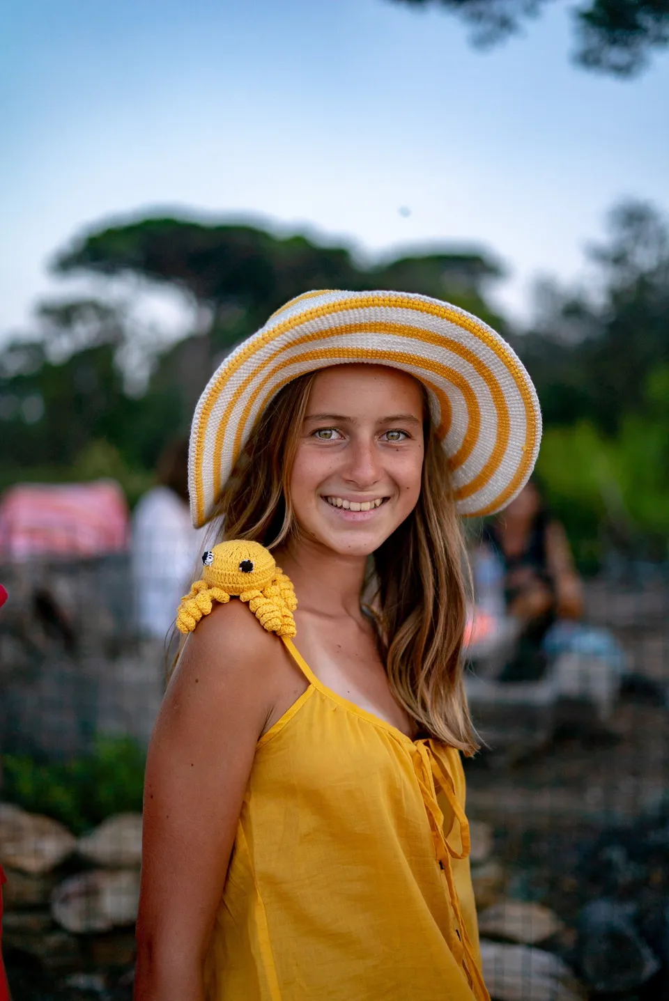 Enfant souriante en blouse jaune, chapeau de crochet rayé jaune-blanc et poulpe en crochet sur l'épaule à Porquerolles.
