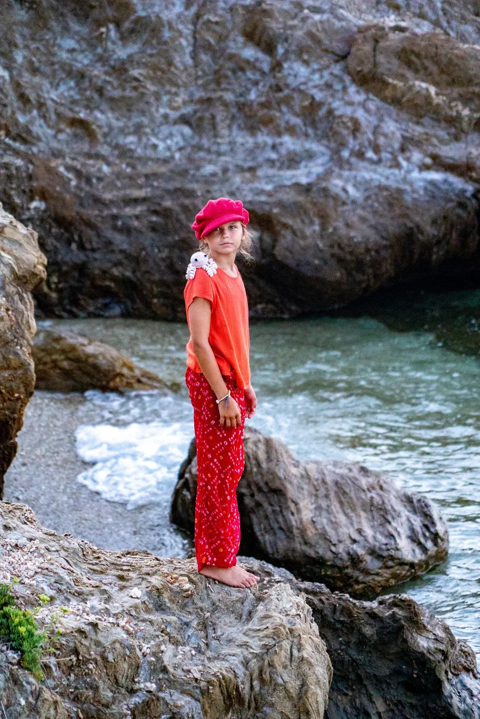Enfant en casquette rouge et pantalon imprimé, pieuvre crochetée sur l'épaule, sur les rochers à Porquerolles.