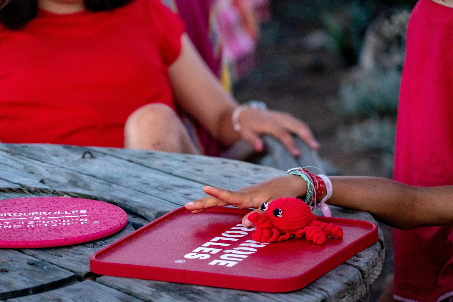 Mains d'enfants autour d'un crabe au crochet rouge posé sur un plateau marqué Porquerolles, table de bois.