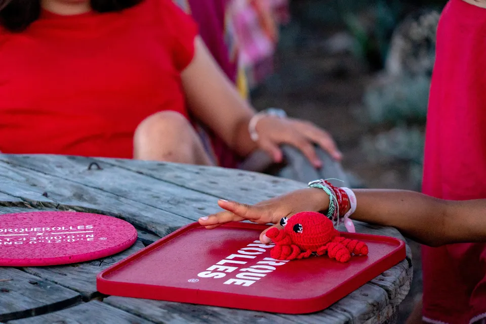 Mains d'enfants autour d'un crabe au crochet rouge posé sur un plateau marqué Porquerolles, table de bois.