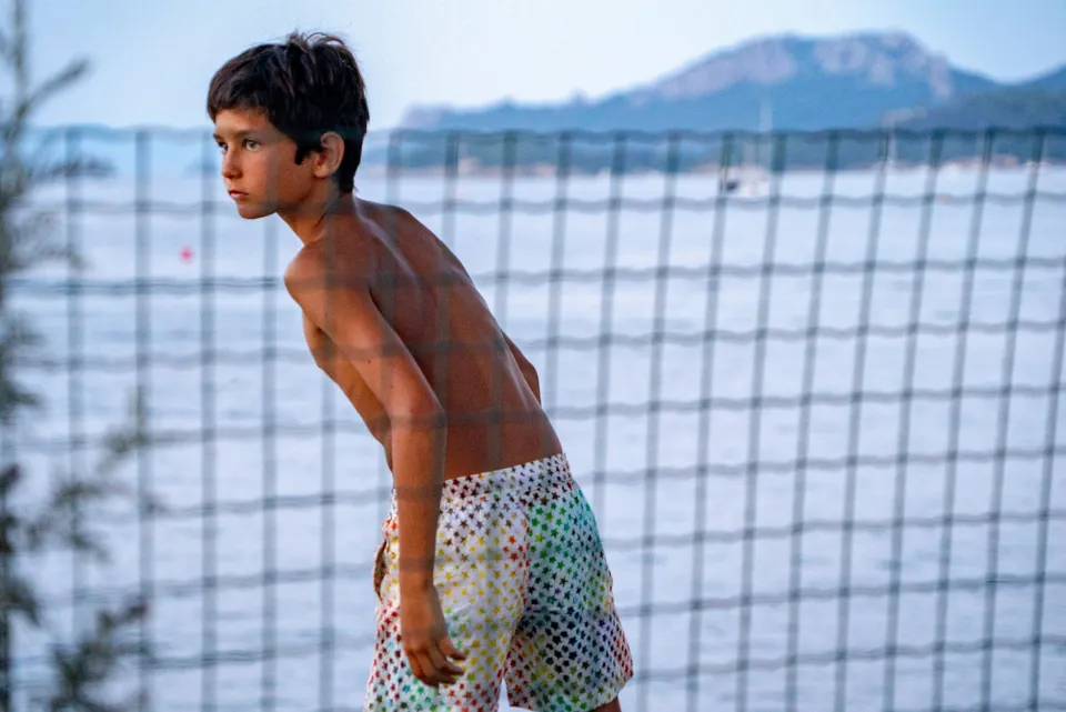 Garçon torse nu en maillot à étoiles, de profil derrière un grillage face à la mer à Porquerolles.