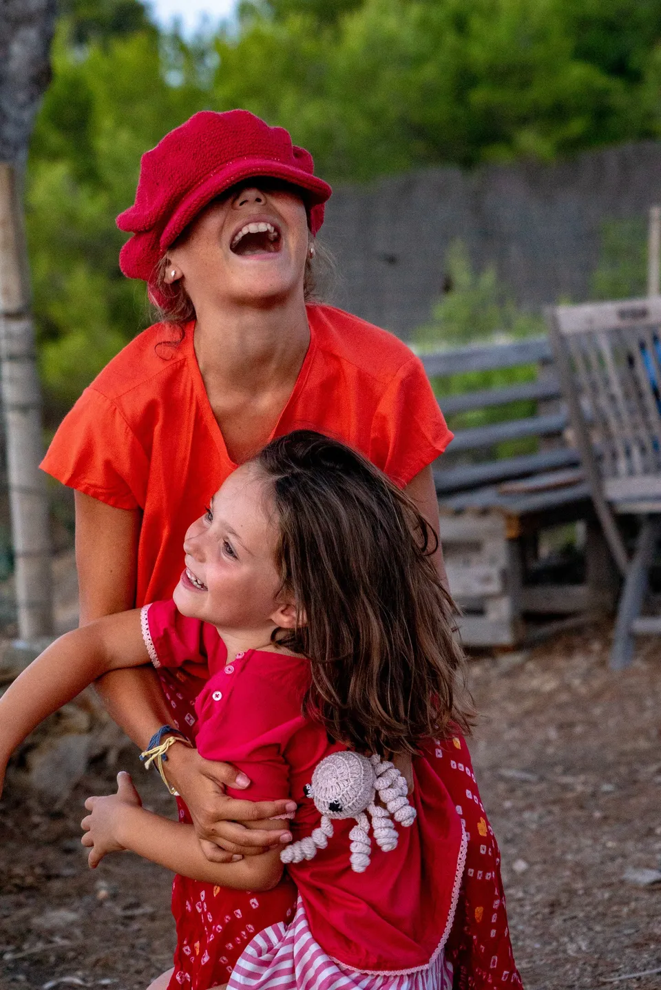 Femme en haut orange et casquette rouge riant, enfant en robe rouge dans ses bras avec une poupée au crochet, à Porquerolles.