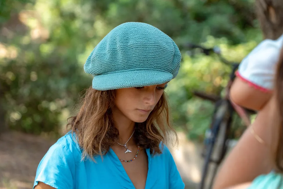 Enfant en casquette turquoise au crochet et chemise bleue, regard baissé, dans le maquis à Porquerolles.