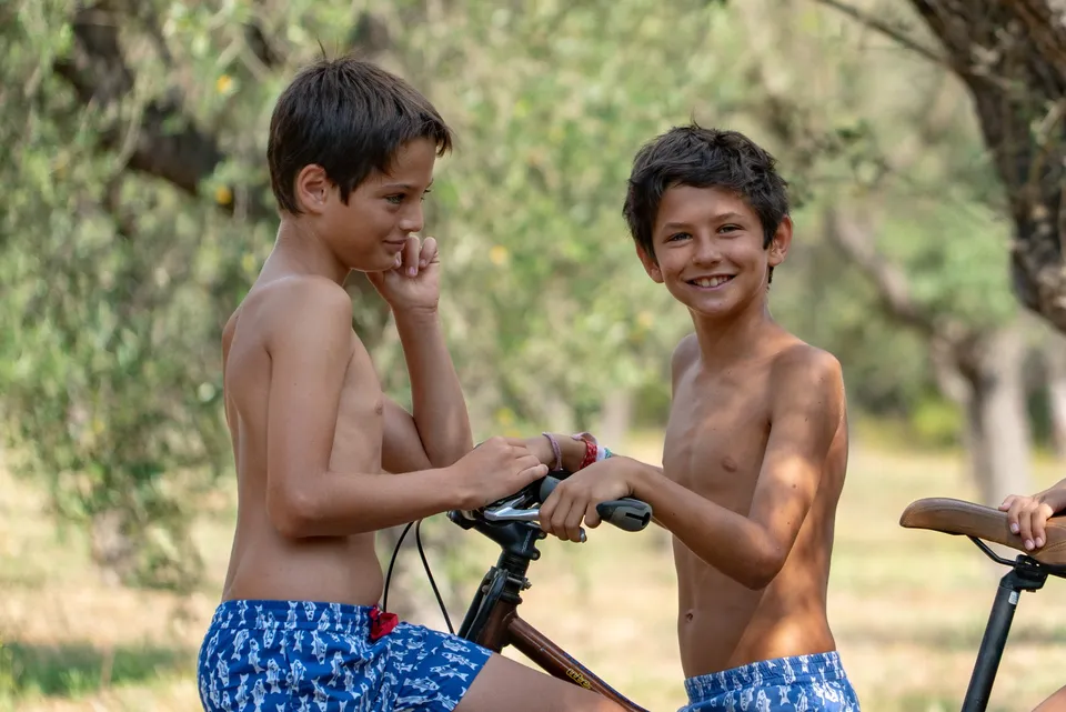 Deux garçons torse nu en maillots bleus à motifs sur des vélos dans une oliveraie ensoleillée à Porquerolles.