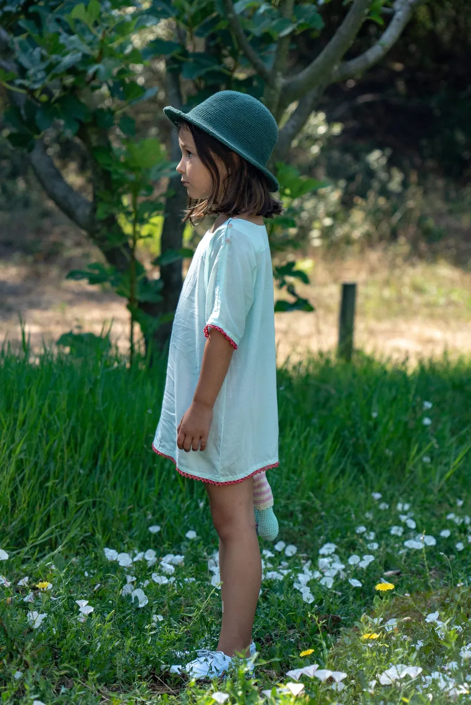 Enfant de profil en tunique menthe ourlée de rose et chapeau de crochet vert, jardin fleuri à Porquerolles.