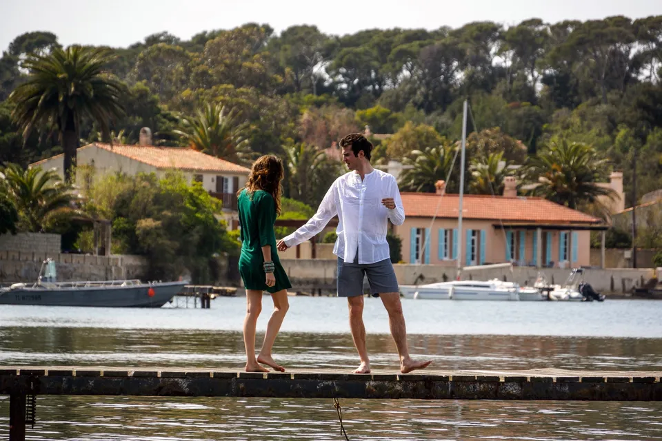 Une femme en robe verte et un homme en chemise blanche marchent pieds nus sur une digue au bord de l’eau.