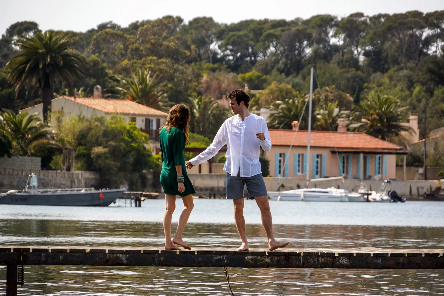 Une femme en robe verte et un homme en chemise blanche marchent pieds nus sur une digue au bord de l’eau.