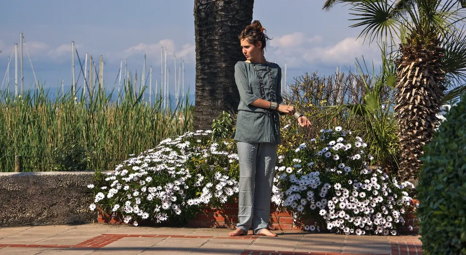 Femme en tunique et pantalon gris-vert debout parmi des marguerites et des palmiers près du port.