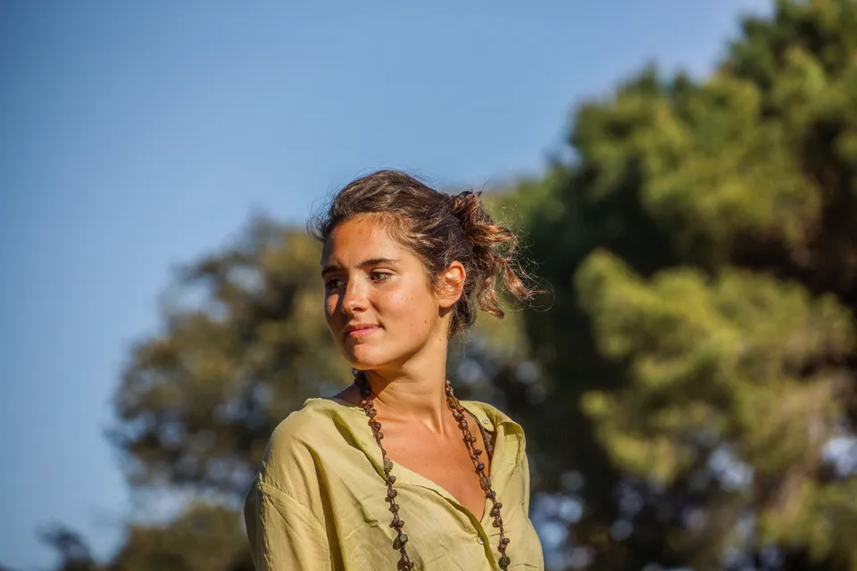 Portrait d’une femme en chemise jaune pâle portant un long collier, devant des arbres.
