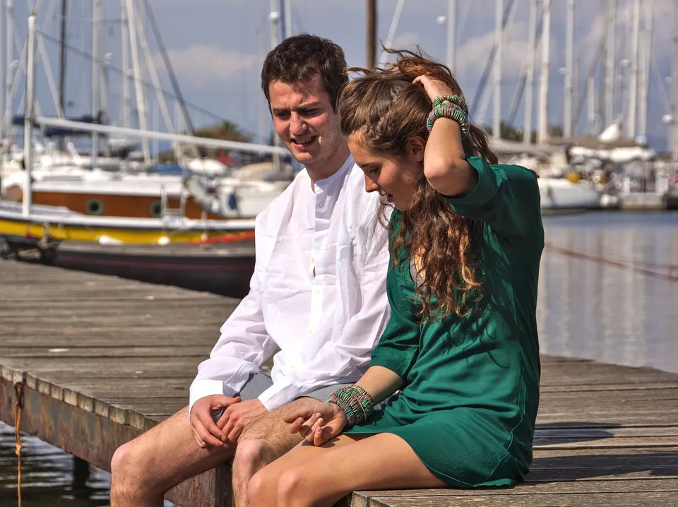 Une femme en robe verte et un homme en chemise blanche sont assis sur un ponton devant les voiliers.