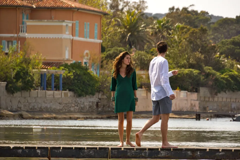 Une femme en robe verte et un homme en chemise blanche marchent au bord de l’eau devant une maison ocre.