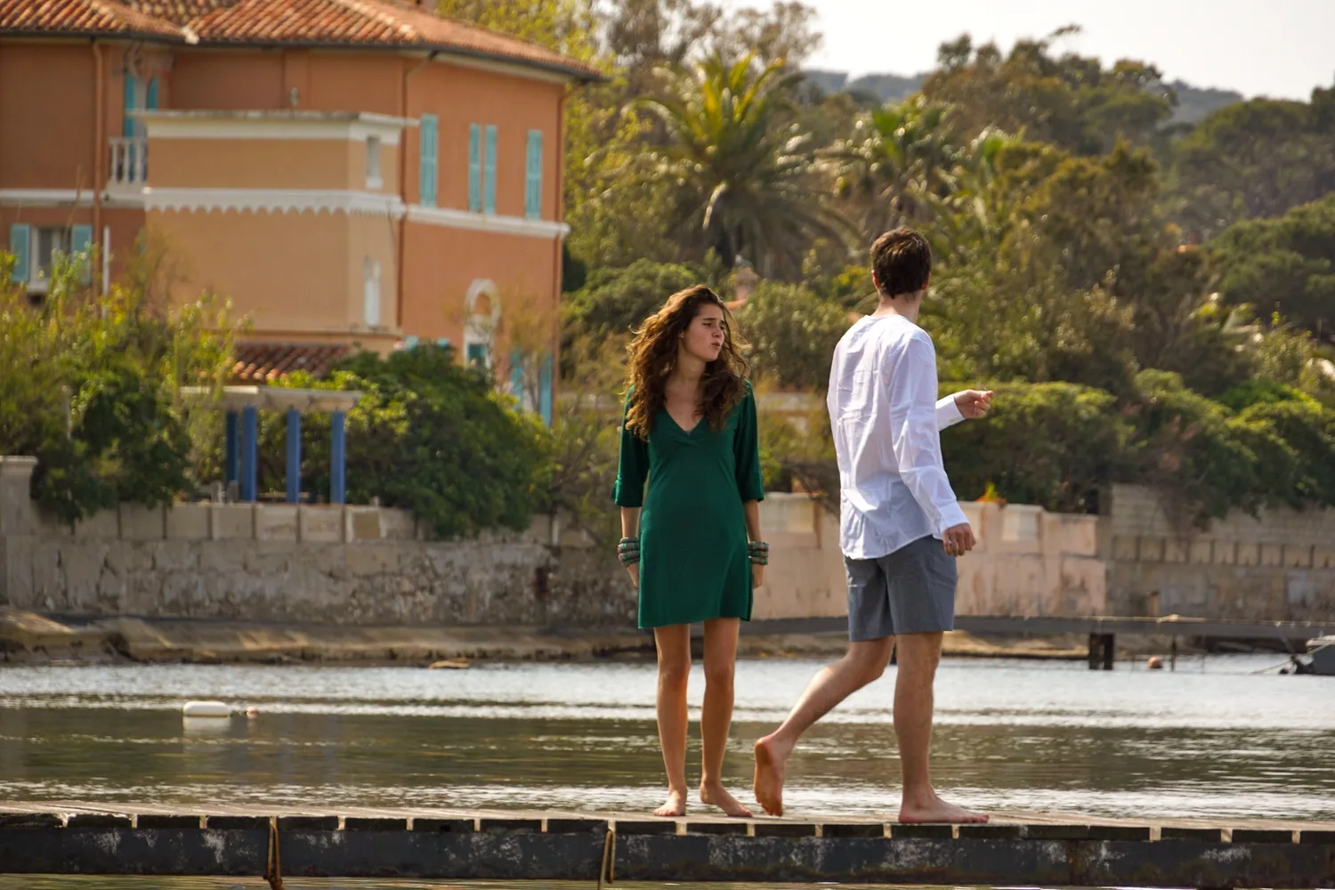Une femme en robe verte et un homme en chemise blanche marchent au bord de l’eau devant une maison ocre.