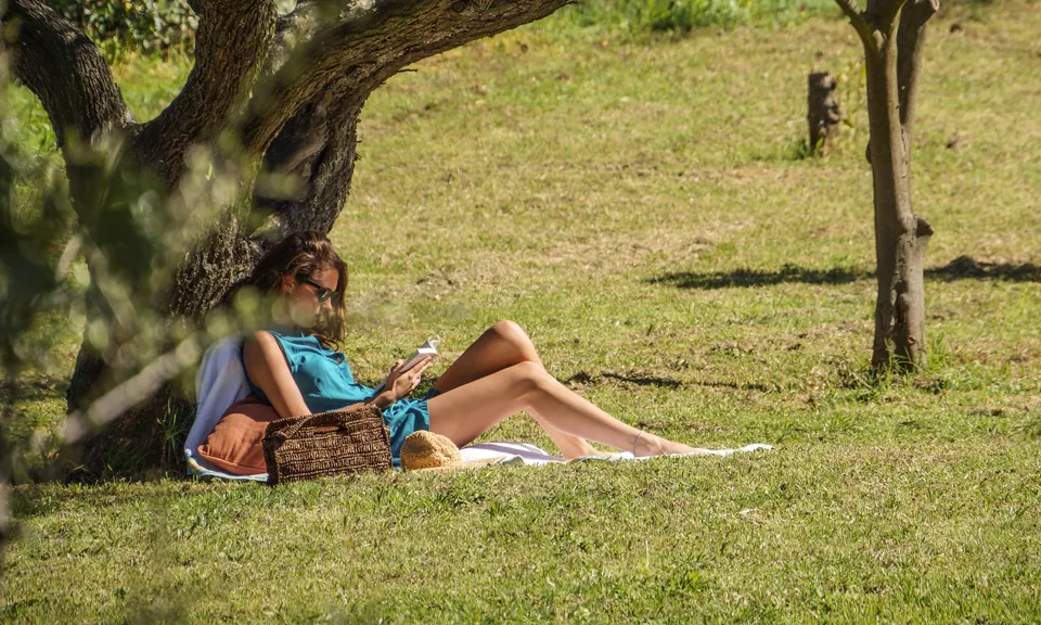 Femme en robe bleue allongée dans l’herbe sous un arbre, avec un chapeau et un sac tressé.