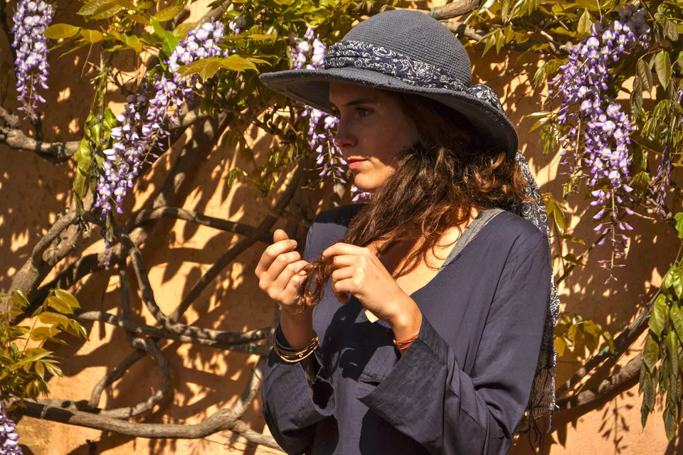 Portrait d’une femme en tunique bleu nuit et chapeau sombre, devant des fleurs violettes.