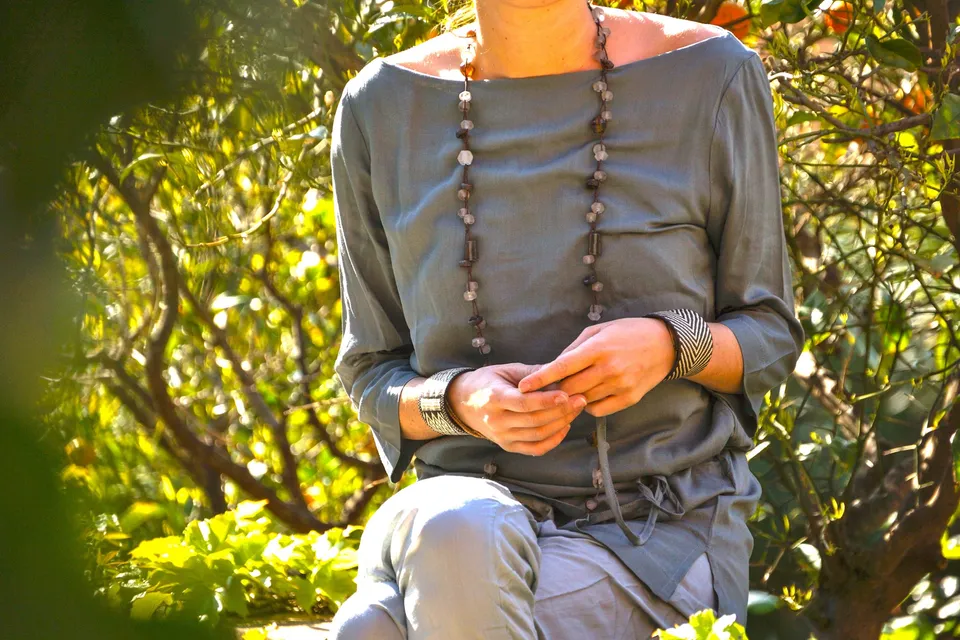 Détail d’une tunique gris-vert, d’un pantalon clair et d’un long collier parmi les feuillages.