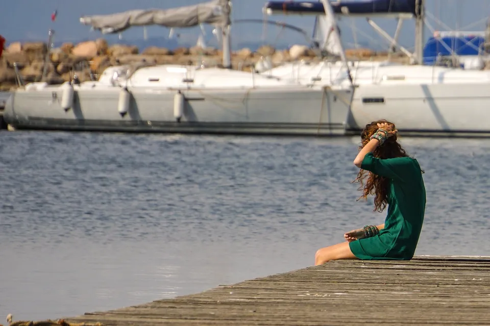 Femme en robe verte assise au bout d’un ponton, face aux bateaux du port.