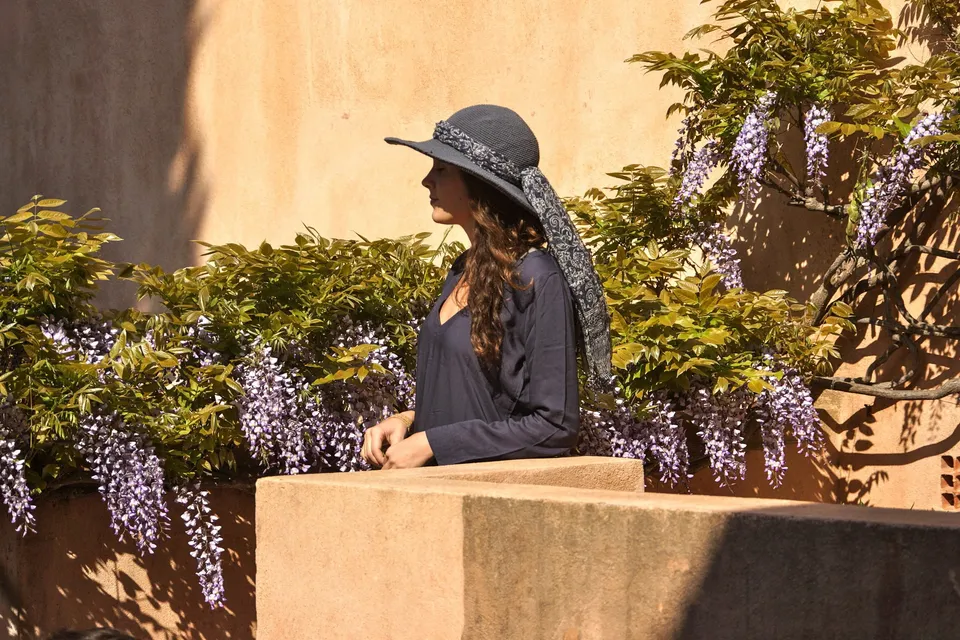 Femme en tunique sombre et chapeau sous une glycine dans un jardin à Porquerolles.