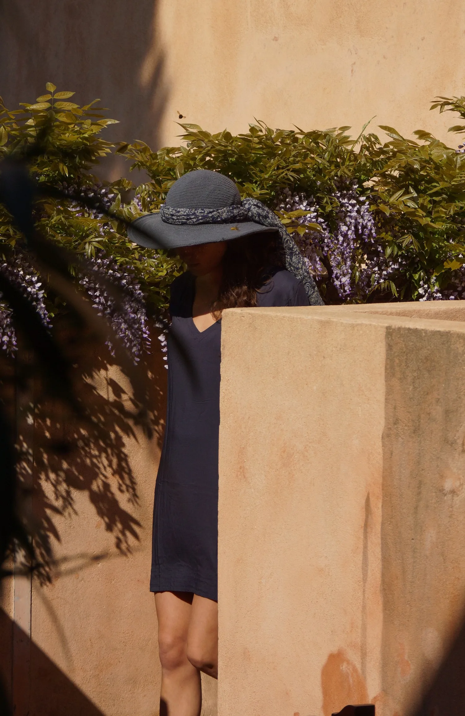 Femme en tunique sombre et chapeau sous une glycine dans un jardin à Porquerolles.
