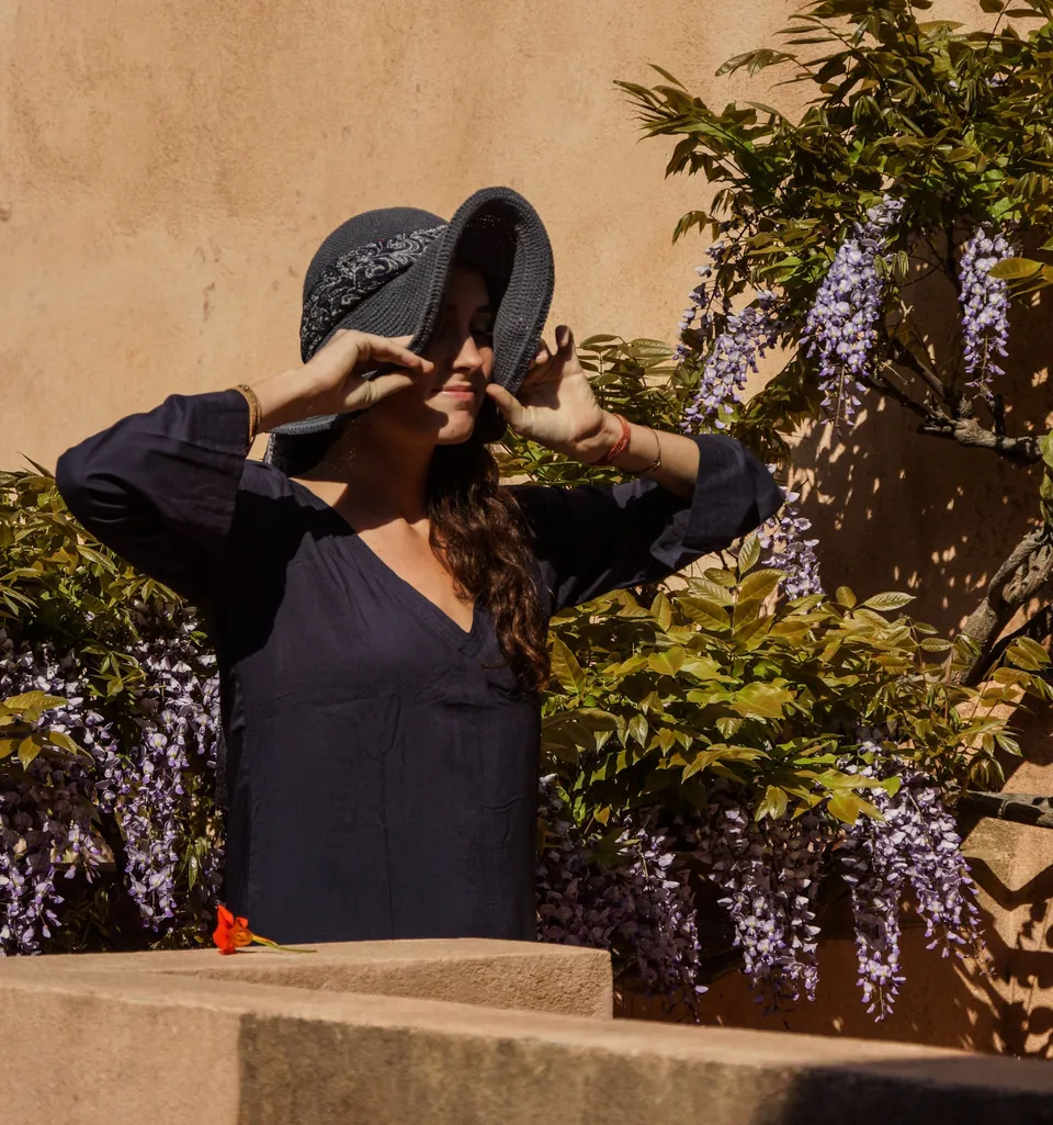Femme en tunique sombre et chapeau sous une glycine dans un jardin à Porquerolles.
