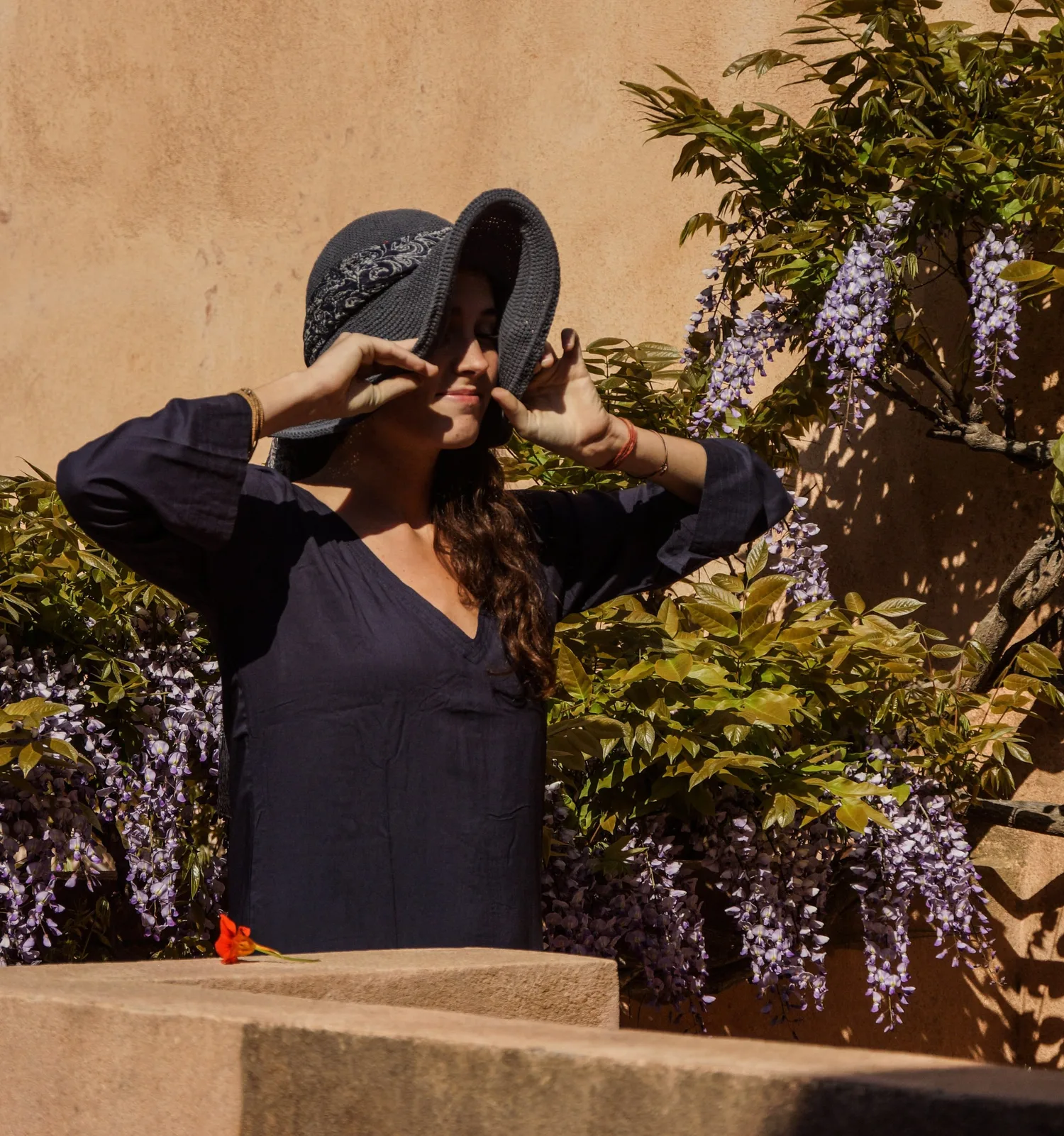 Femme en tunique sombre et chapeau sous une glycine dans un jardin à Porquerolles.