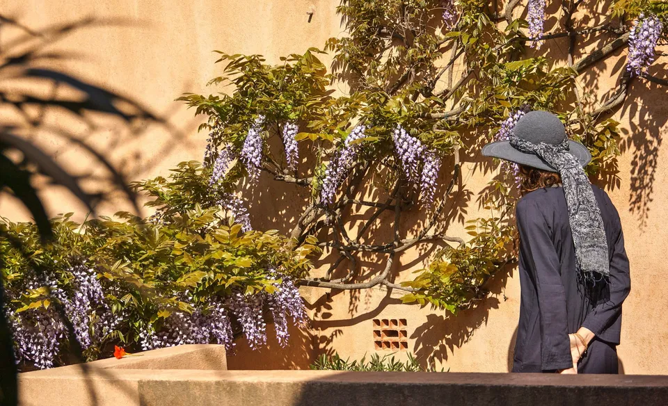 Femme en tunique sombre et chapeau sous une glycine dans un jardin à Porquerolles.