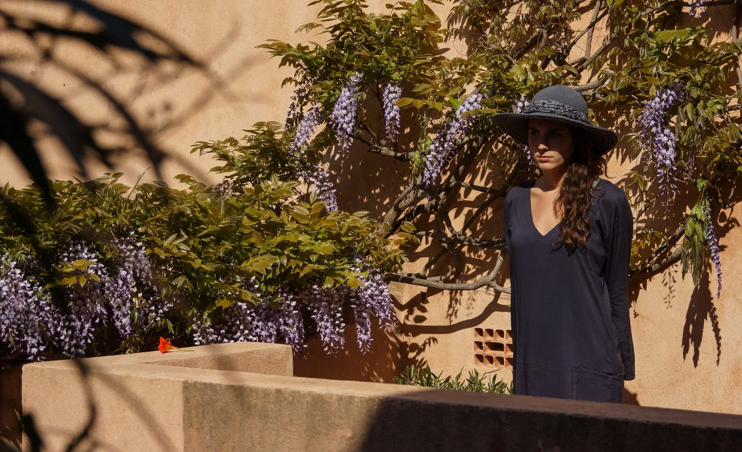 Femme en tunique sombre et chapeau sous une glycine dans un jardin à Porquerolles.