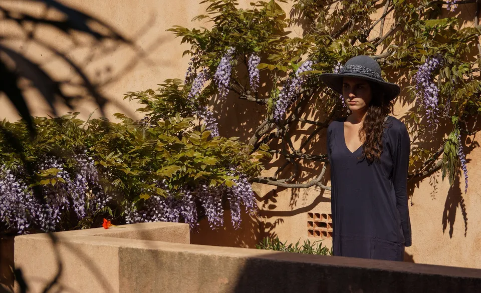 Femme en tunique sombre et chapeau sous une glycine dans un jardin à Porquerolles.