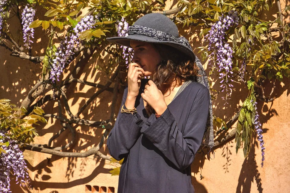 Femme en tunique sombre et chapeau sous une glycine dans un jardin à Porquerolles.