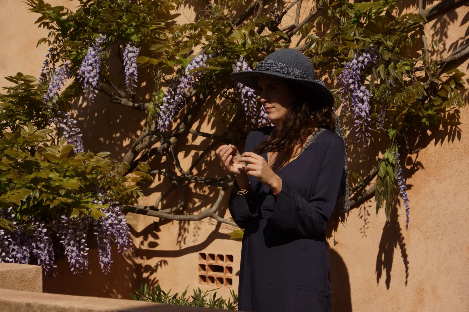Femme en tunique sombre et chapeau sous une glycine dans un jardin à Porquerolles.