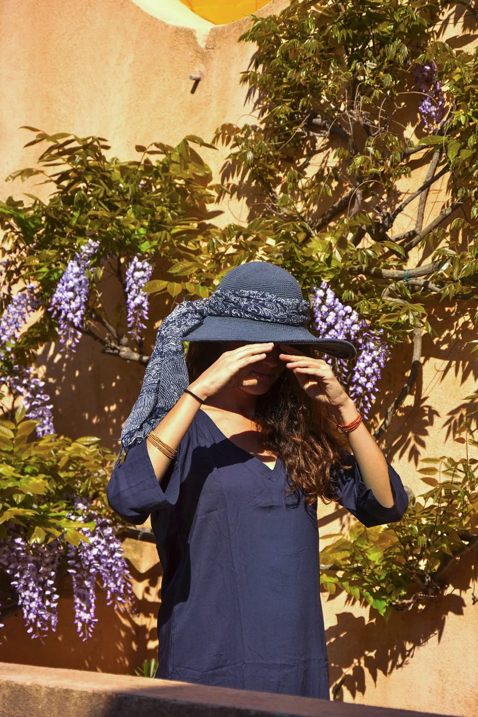 Femme en tunique sombre et chapeau sous une glycine dans un jardin à Porquerolles.