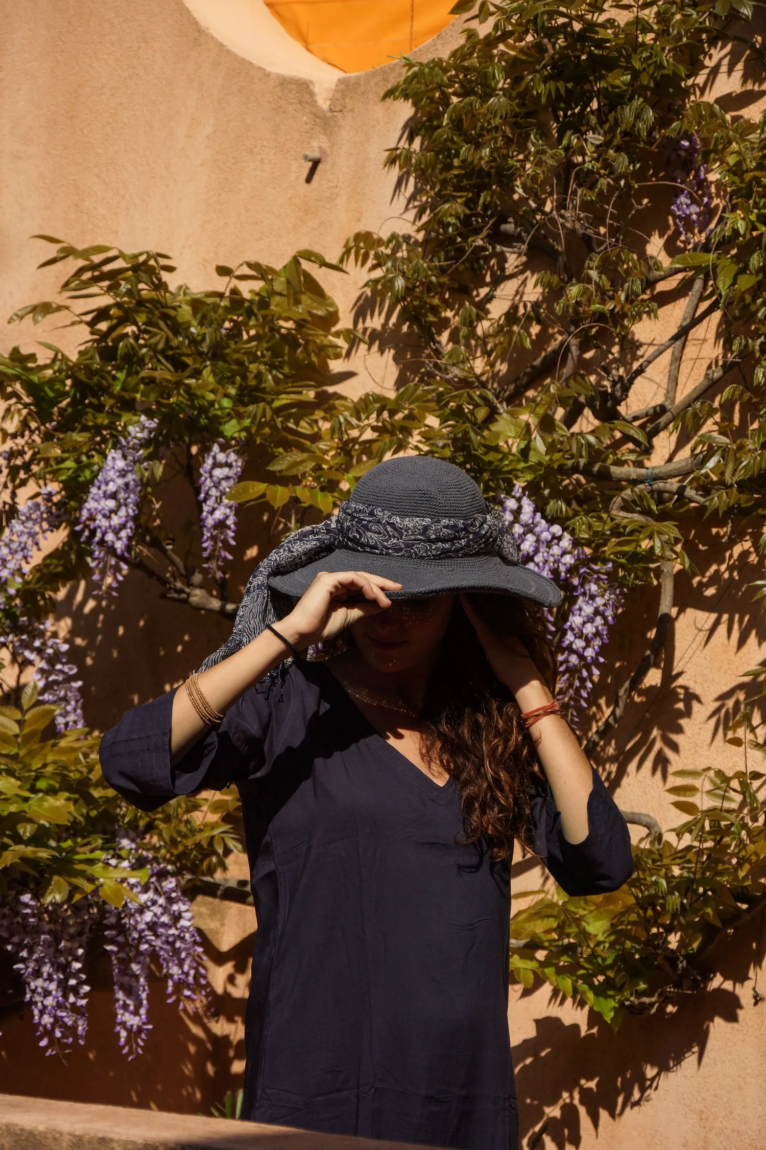 Femme en tunique sombre et chapeau sous une glycine dans un jardin à Porquerolles.