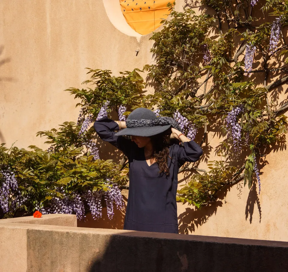 Femme en tunique sombre et chapeau sous une glycine dans un jardin à Porquerolles.