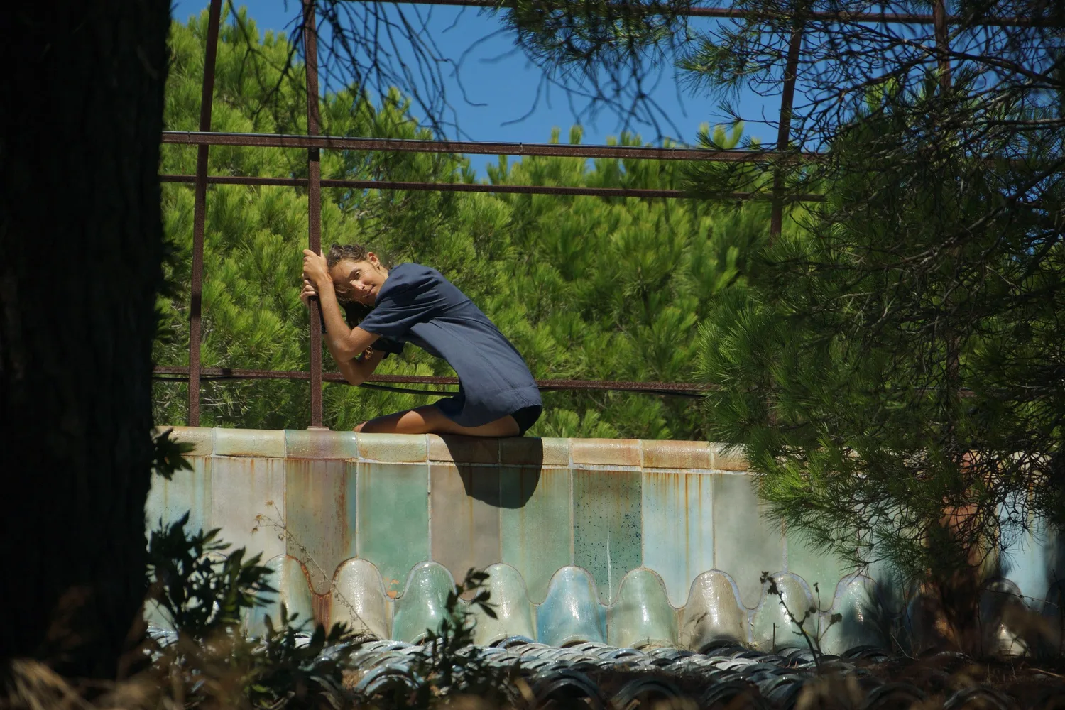Femme en robe-chemise de coton bleu agrippée à une pergola de fer rouillé au-dessus d'un bassin, sous les pins à Porquerolles.