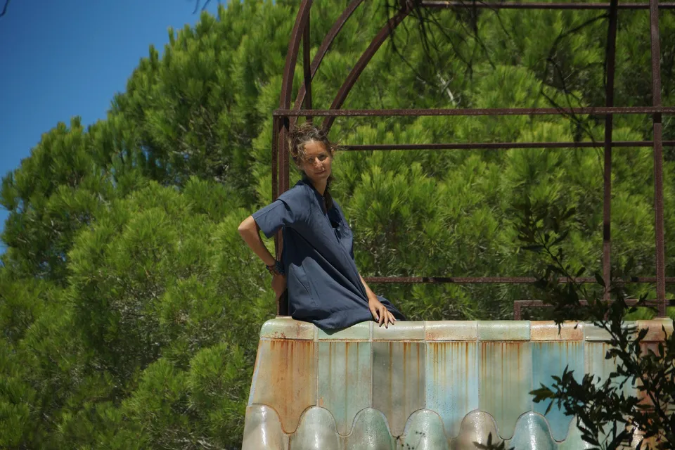 Femme en robe-chemise de coton bleu debout derrière un bassin de pierre, main sur la hanche, ciel bleu et pins à Porquerolles.