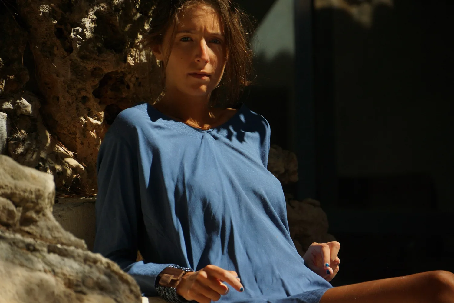 Portrait d'une femme en robe de coton bleu à manches longues, bracelet de perles au poignet, adossée à la roche à Porquerolles.