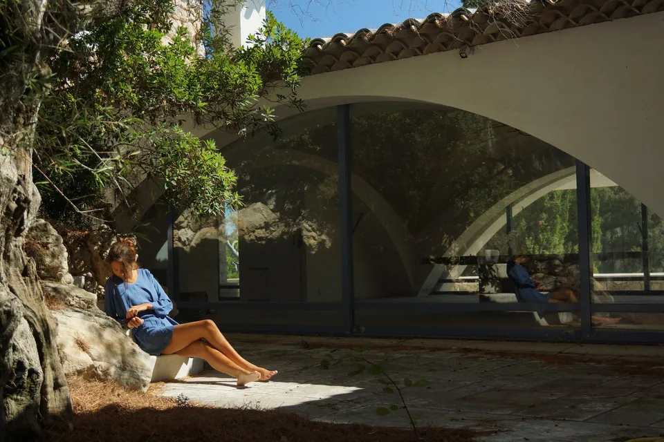Femme en robe bleue assise sous un vieil olivier dans la cour d'une maison à arcades vitrées à Porquerolles.