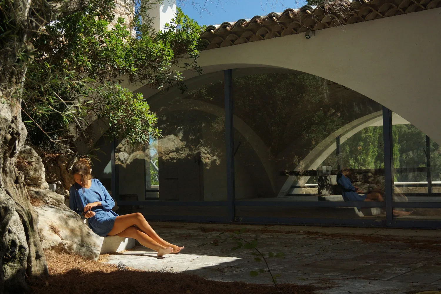Femme en robe bleue assise sous un vieil olivier dans la cour d'une maison à arcades vitrées à Porquerolles.
