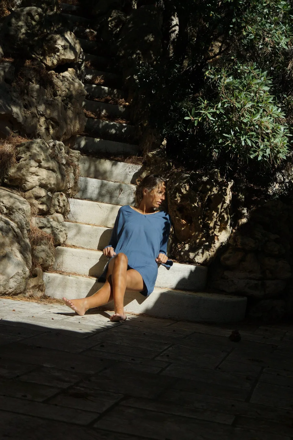 Femme en robe de coton bleu assise pieds nus sur un escalier de pierre taillé dans la roche, tache de soleil, à Porquerolles.
