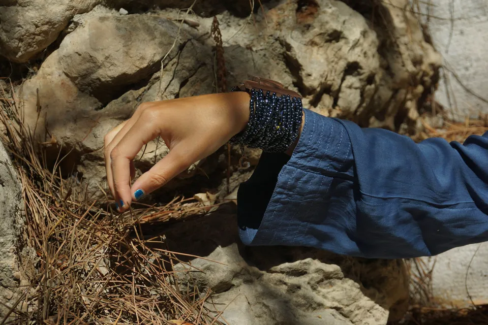 Détail d'une main aux ongles bleus effleurant des aiguilles de pin, manchette de perles et manche de coton bleu, à Porquerolles.
