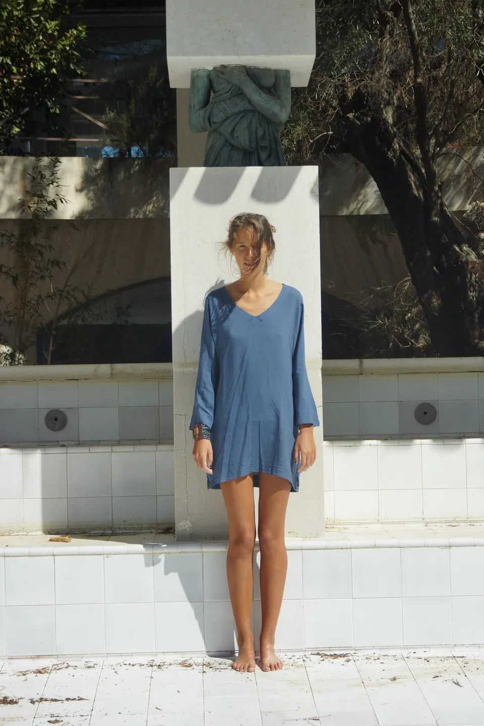 Femme en robe de coton bleu debout pieds nus contre un pilier blanc, devant un bassin carrelé et une sculpture, à Porquerolles.